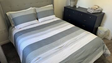 A neatly made bed with a striped bedspread, a grey upholstered headboard, a bedside table with a lamp, and a small trash bin.