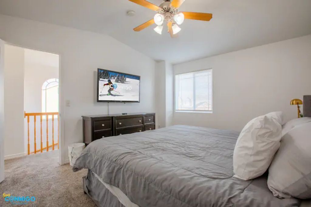 A bedroom with a bed, a ceiling fan, a TV, a window, and a door leading to another room.