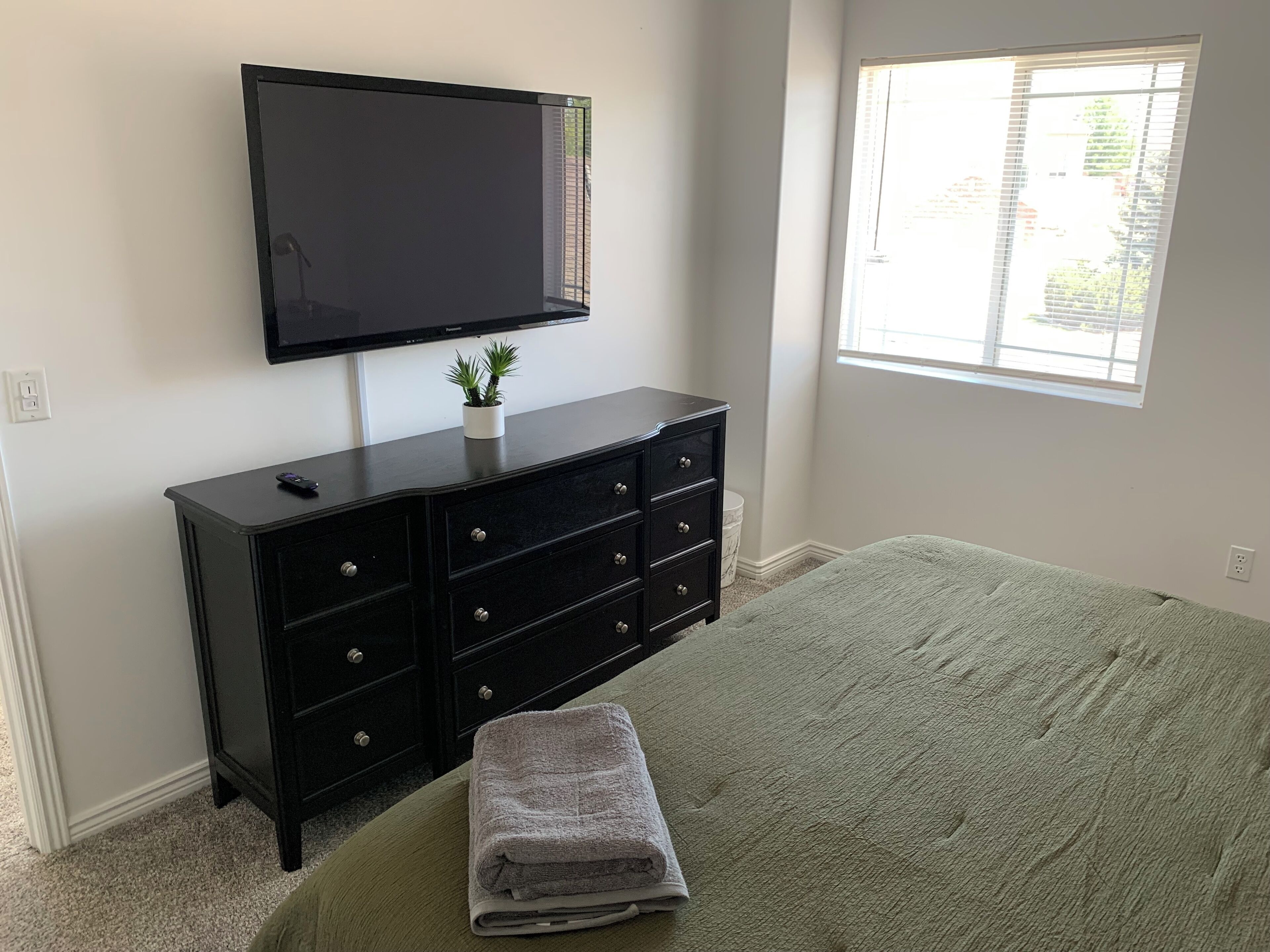 Un dormitorio con cama, cómoda, televisión montada en la pared y ventana con persianas.