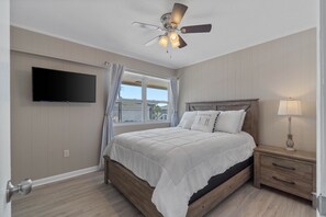 Une chambre à coucher comprenant un lit, une table de chevet, une lampe, un ventilateur de plafond et un téléviseur fixé au mur.