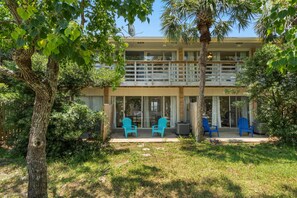 Un bâtiment à deux étages doté d’un balcon, d’un espace extérieur aménagé avec des chaises bleues, et d’un arbre au premier plan.