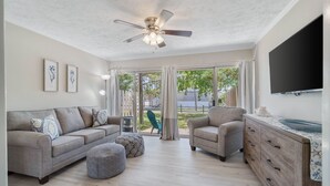 Un salón amplio con ventilador de techo, un televisor grande y vistas al exterior a través de amplias ventanas.