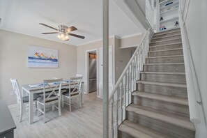 Un coin repas comprenant une table et des chaises, un ventilateur de plafond et un escalier avec des marches en bois et des rampes blanches.