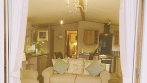 Una sala de estar con un sofá color beige, dos sillones a juego y un reposapiés blanco. Un candelabro cuelga del techo, y se ve una cocina al fondo.