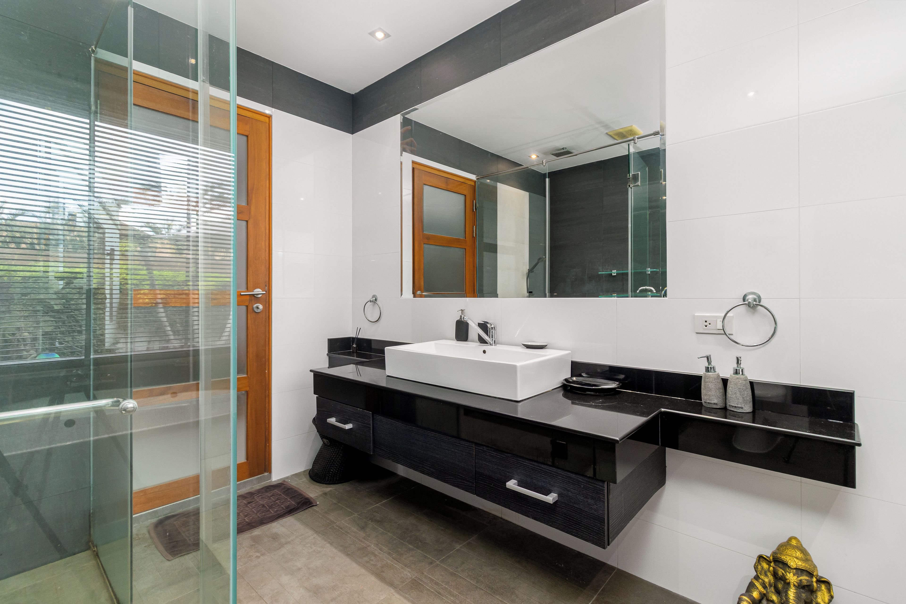 Un baño moderno con cabina de ducha de vidrio, puerta de madera, lavabo blanco y mueble de tocador negro.