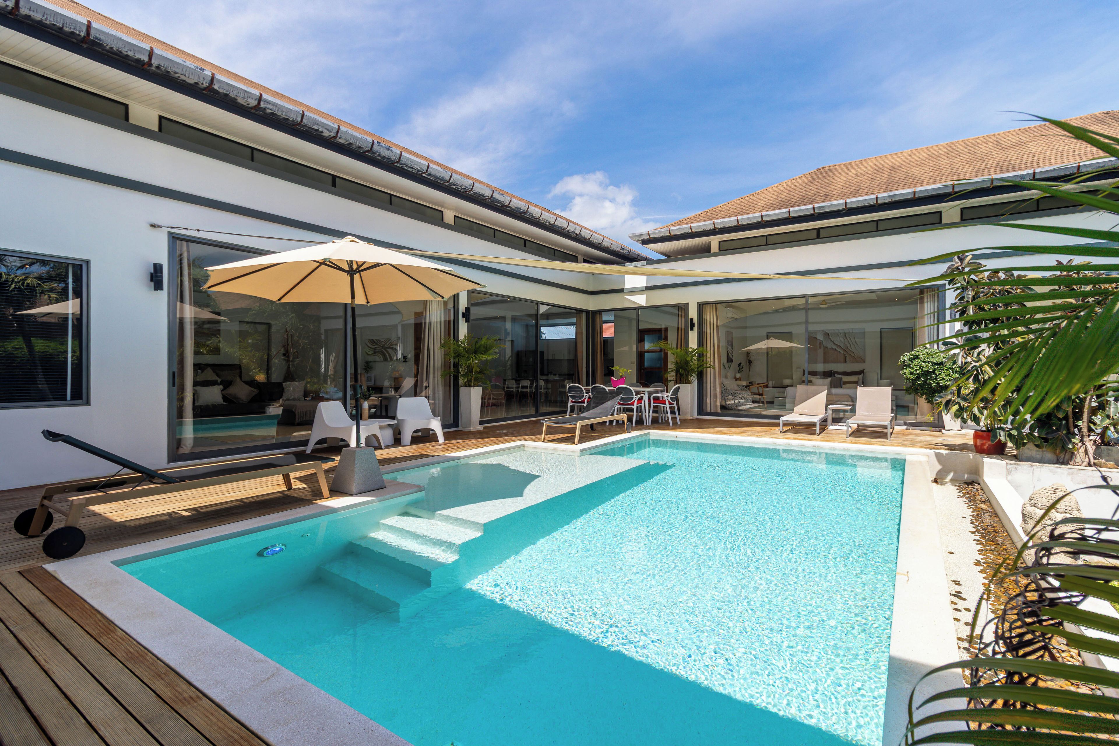 A modern outdoor pool area with a wooden deck, lounge chairs, and a glass-enclosed patio.