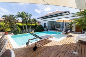 A modern outdoor pool area with two lounge chairs, a wooden deck, and a glass-enclosed patio. - Modern 4BR Pool Villa Virginie Naiharn (Rawai)