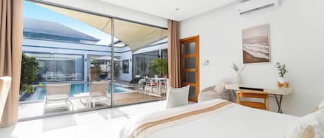 A modern hotel room with a large bed, a desk, a view of a pool and outdoor seating, and a beach scene painting.