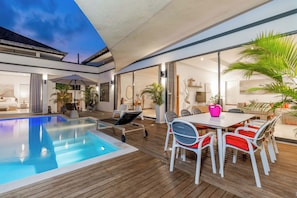 A modern outdoor pool area with a wooden deck, lounge chairs, and a dining table with chairs.