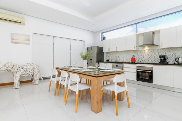 A modern kitchen with white cabinetry, a dining area with a glass table and chairs, and a view of the sky through a window.