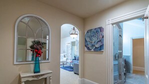 Um corredor com um espelho decorativo, um vaso azul com fita vermelha e um grande quadro floral na parede.