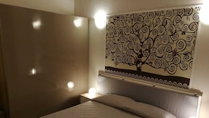 A bedroom with a bed, two bedside tables with lamps, a wall with a tree mural, and a wooden wardrobe.