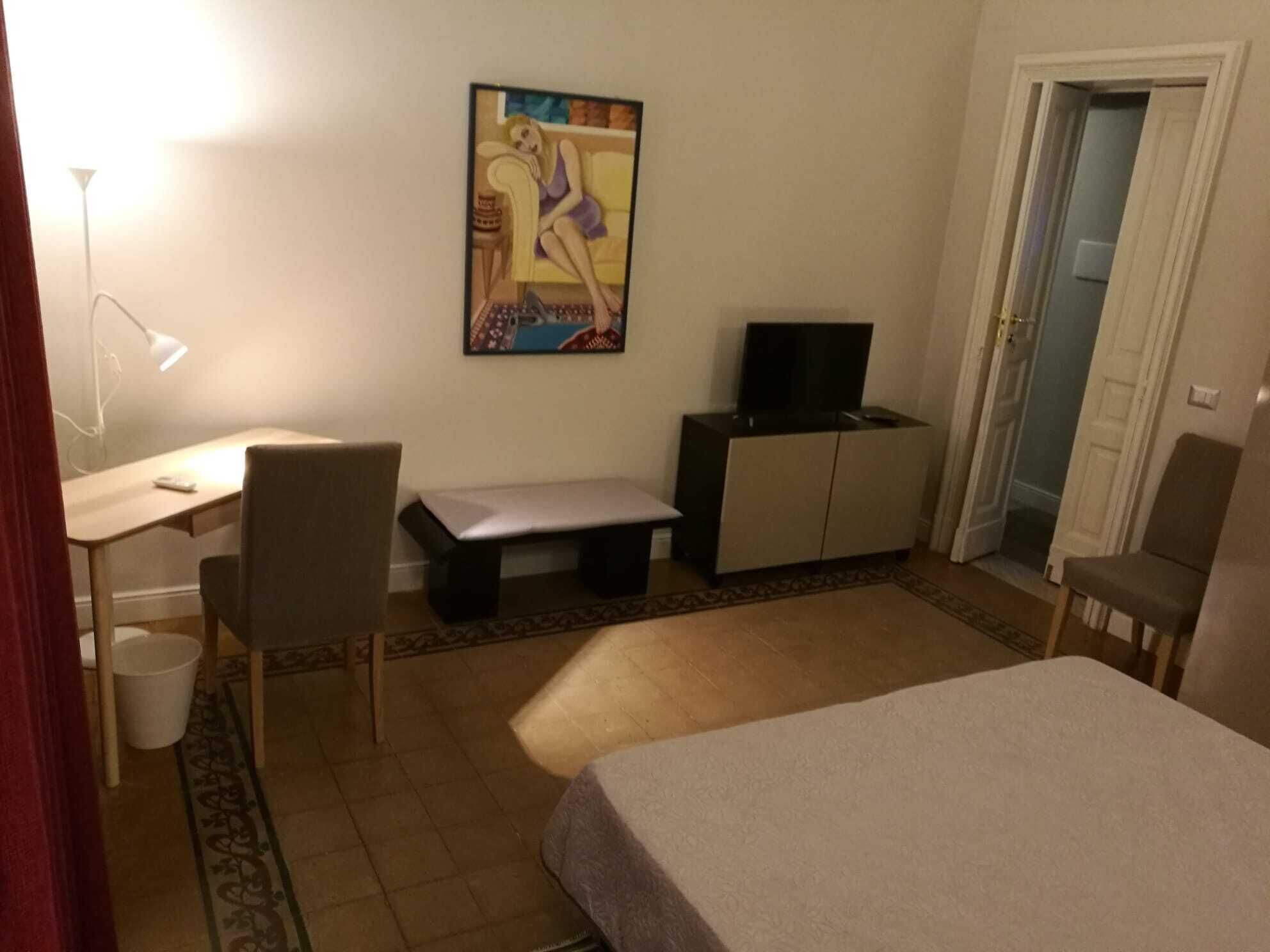 Ett litet, möblerat rum med en säng, ett skrivbord med en stol, en tv på ett skåp och en tavla på väggen.