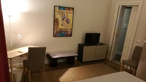 A small, furnished room with a bed, a desk with a chair, a television on a cabinet, and a painting on the wall.