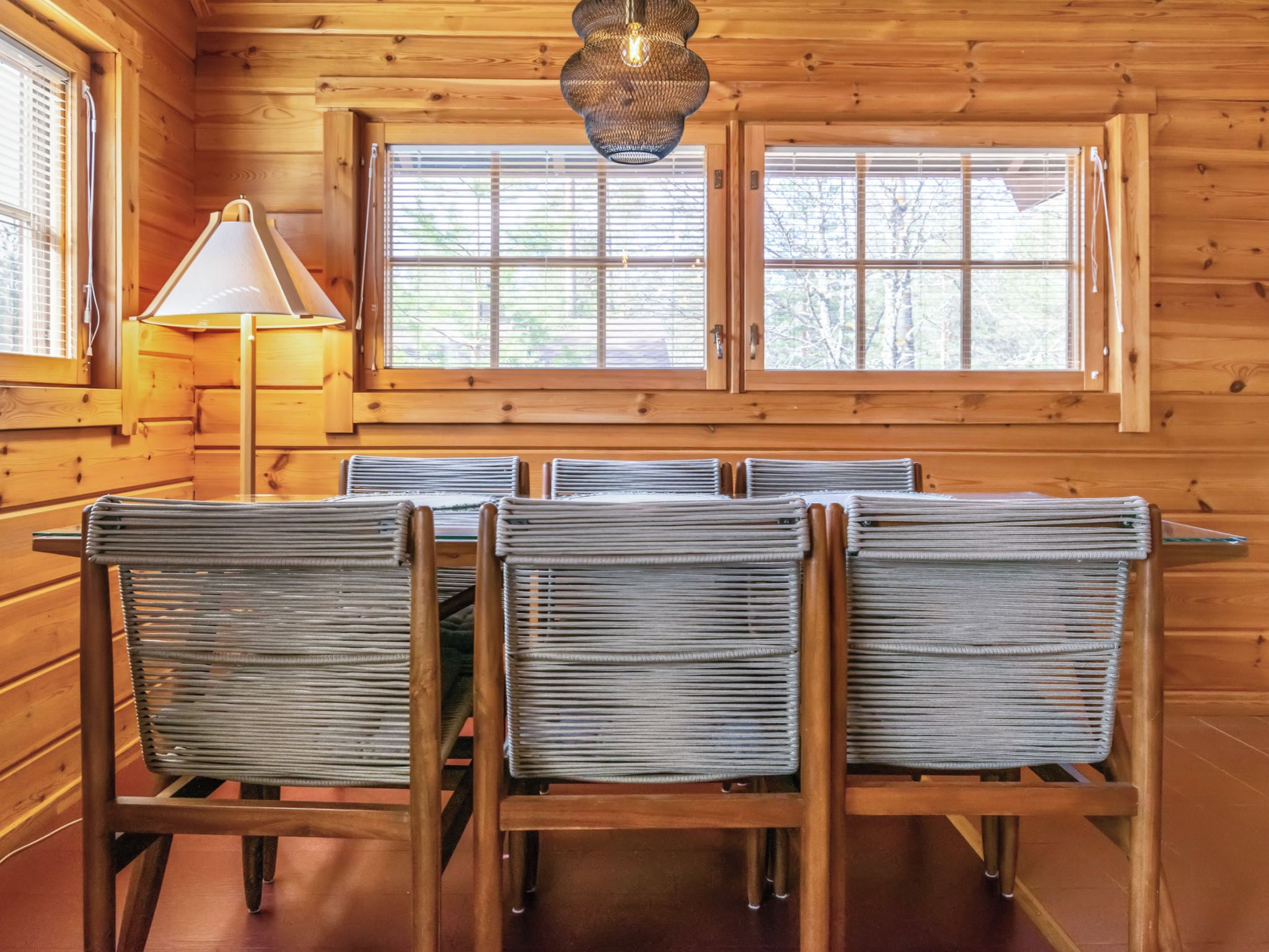 A wooden interior with a dining table and chairs, a lamp, and windows.