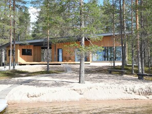 Una cabaña moderna con fachada de madera, ubicada cerca de una playa arenosa y rodeada de árboles.