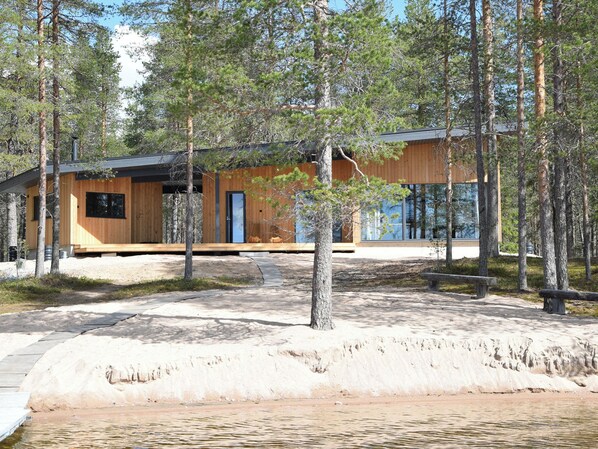 En moderne hytte med træfacade, beliggende nær en sandstrand og omgivet af træer.