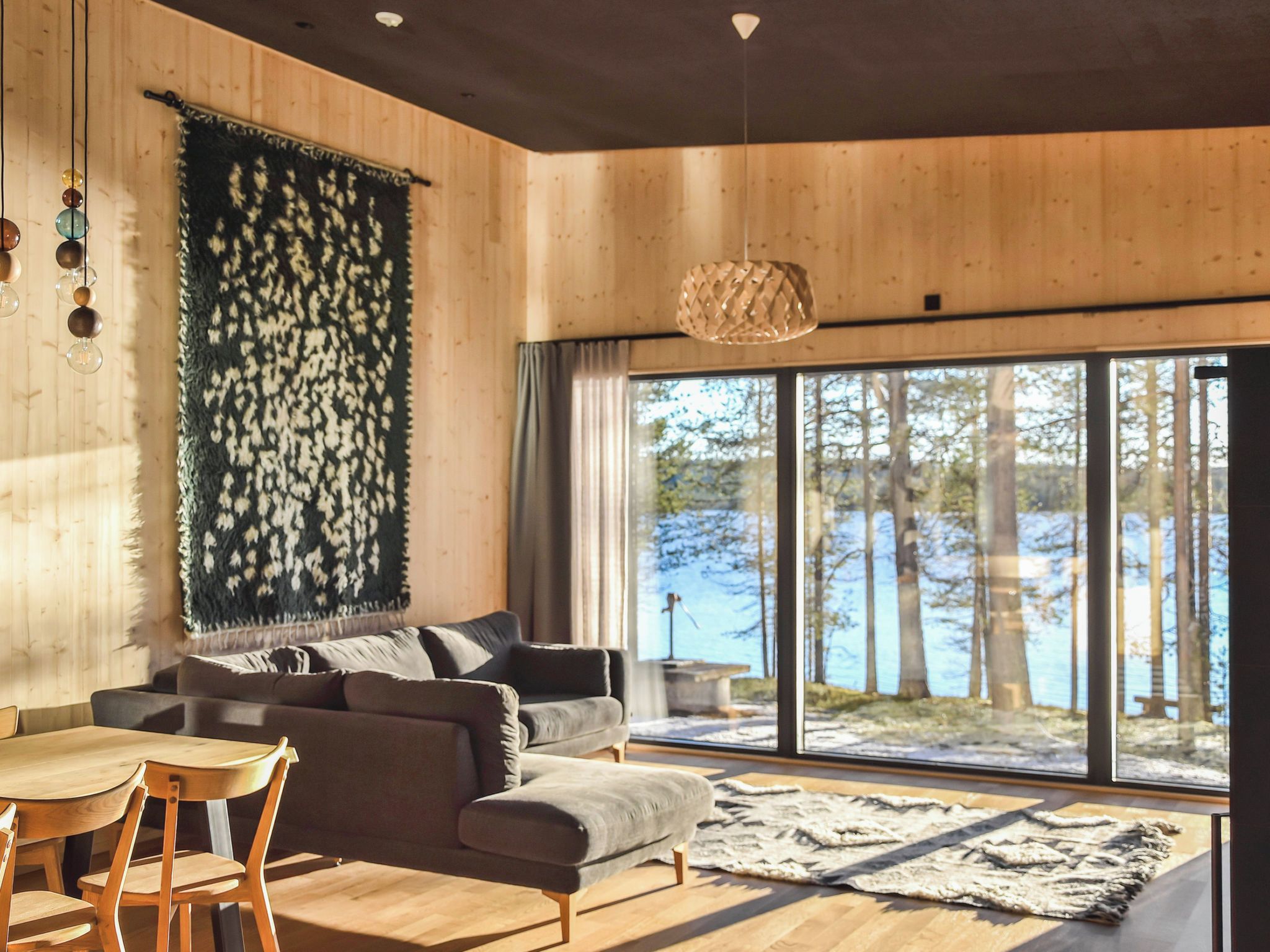 A modern interior with a wooden floor, a sofa, a dining table, and a large window with a view of trees.