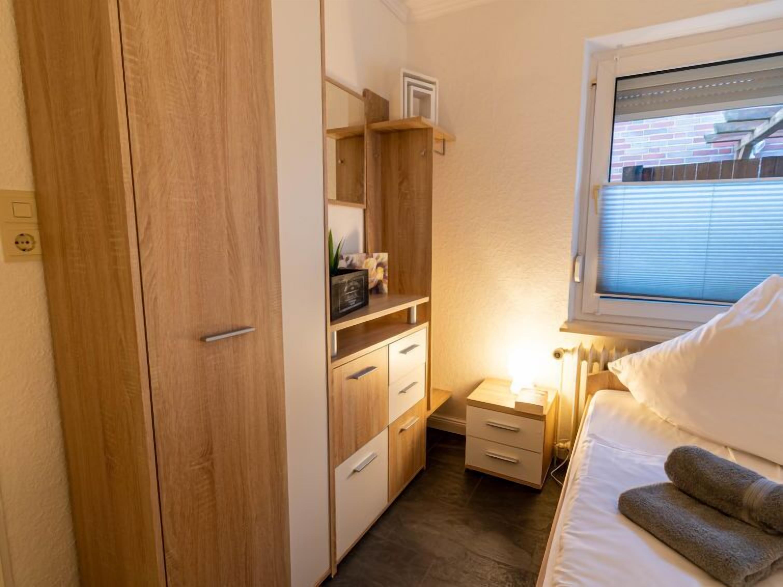 Une chambre à coucher de taille modeste, avec une armoire en bois, une table de chevet avec une lampe, un lit avec du linge de lit blanc et une fenêtre avec des stores.