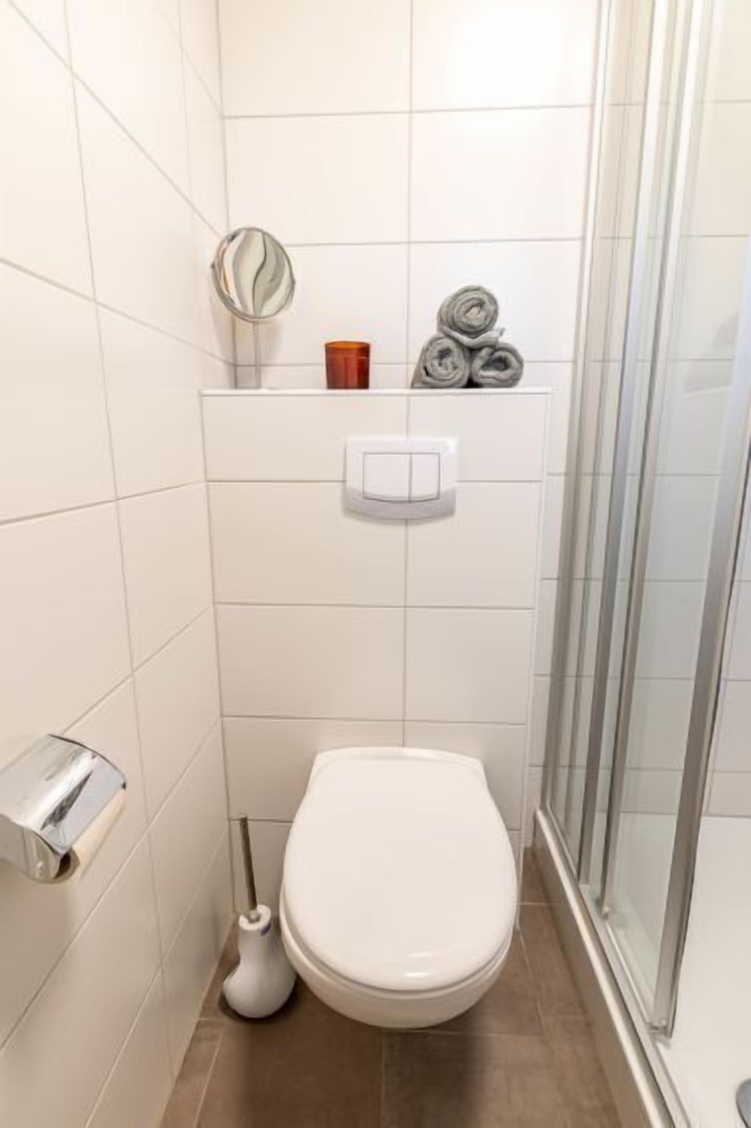 Et lille, moderne badeværelse med hvide fliser, toilet, hårtørrer, toiletpapirholder og bruseområde.