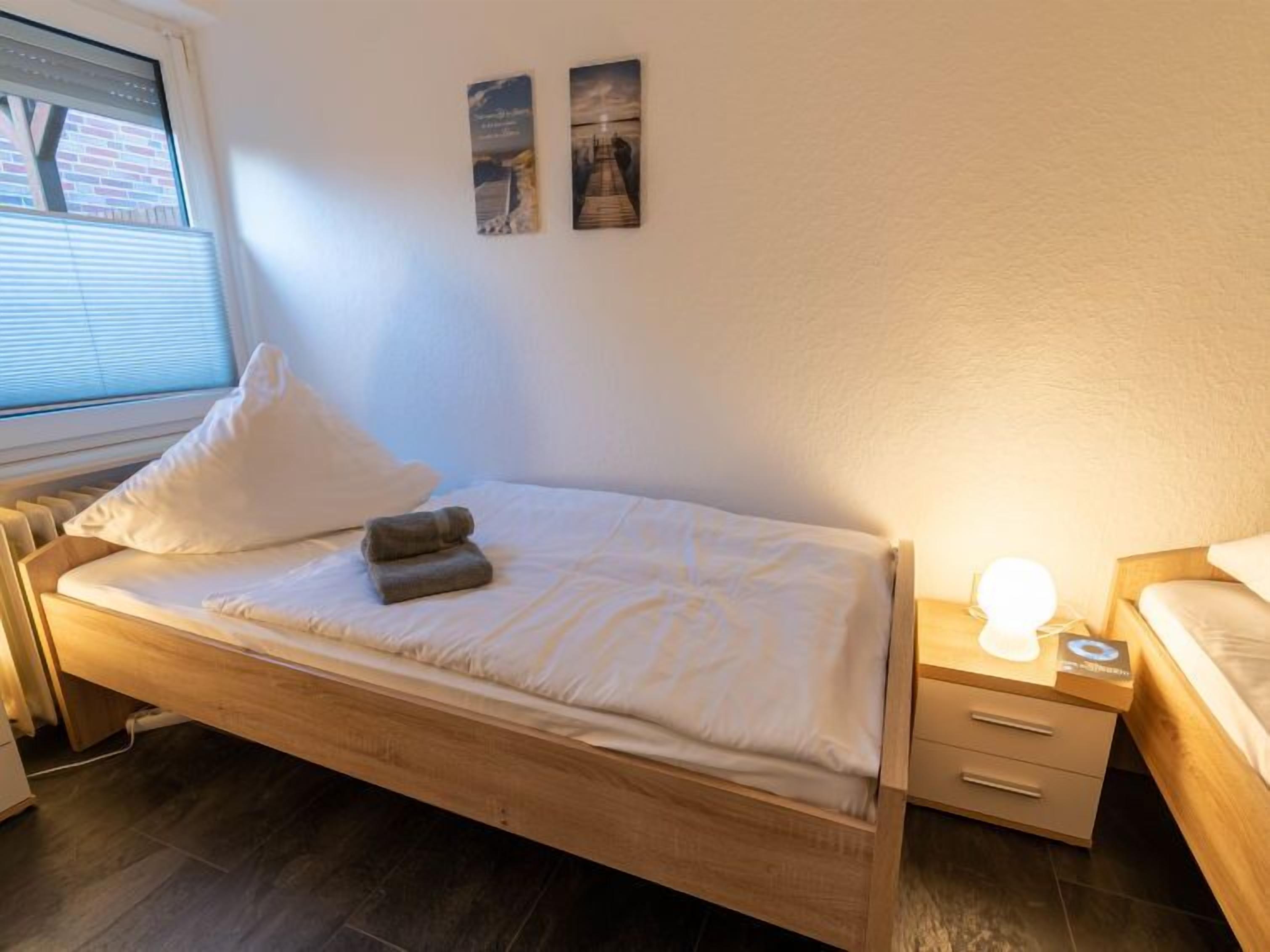 En pænt redt seng med pude og et foldet håndklæde, et natbord med lampe, og et vindue med rullegardiner.