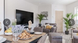 Una sala de estar moderna con un televisor de pantalla plana, asientos cómodos, una zona de comedor con una mesa dispuesta para dos personas y una planta en maceta.