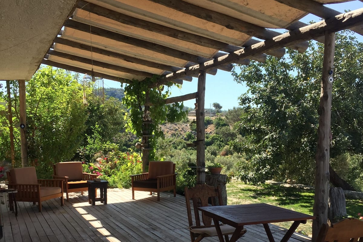 Una terraza de madera con una pérgola, mobiliario exterior y vistas a un paisaje exuberante.