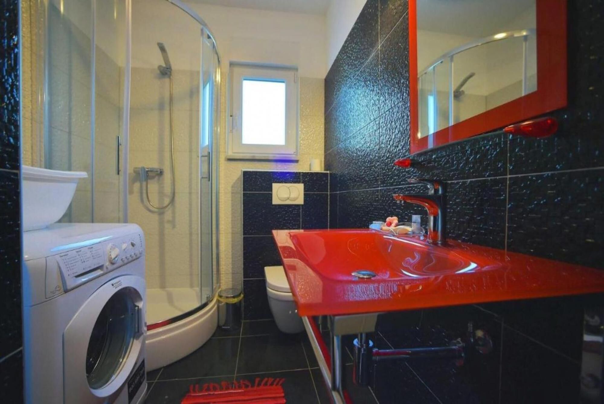 A modern bathroom with a glass shower enclosure, a washing machine, a toilet, and a red sink with a mirror above it.