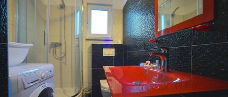 A modern bathroom with a glass shower enclosure, a washing machine, a toilet, and a red sink with a mirror above it.