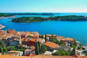 A coastal town with terracotta-roofed buildings, a clear blue sea, and a small island in the distance.