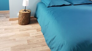 A bedroom with a sloped ceiling, a bed with blue bedding, a round wooden side table with a lamp, and a herringbone-patterned wooden floor.