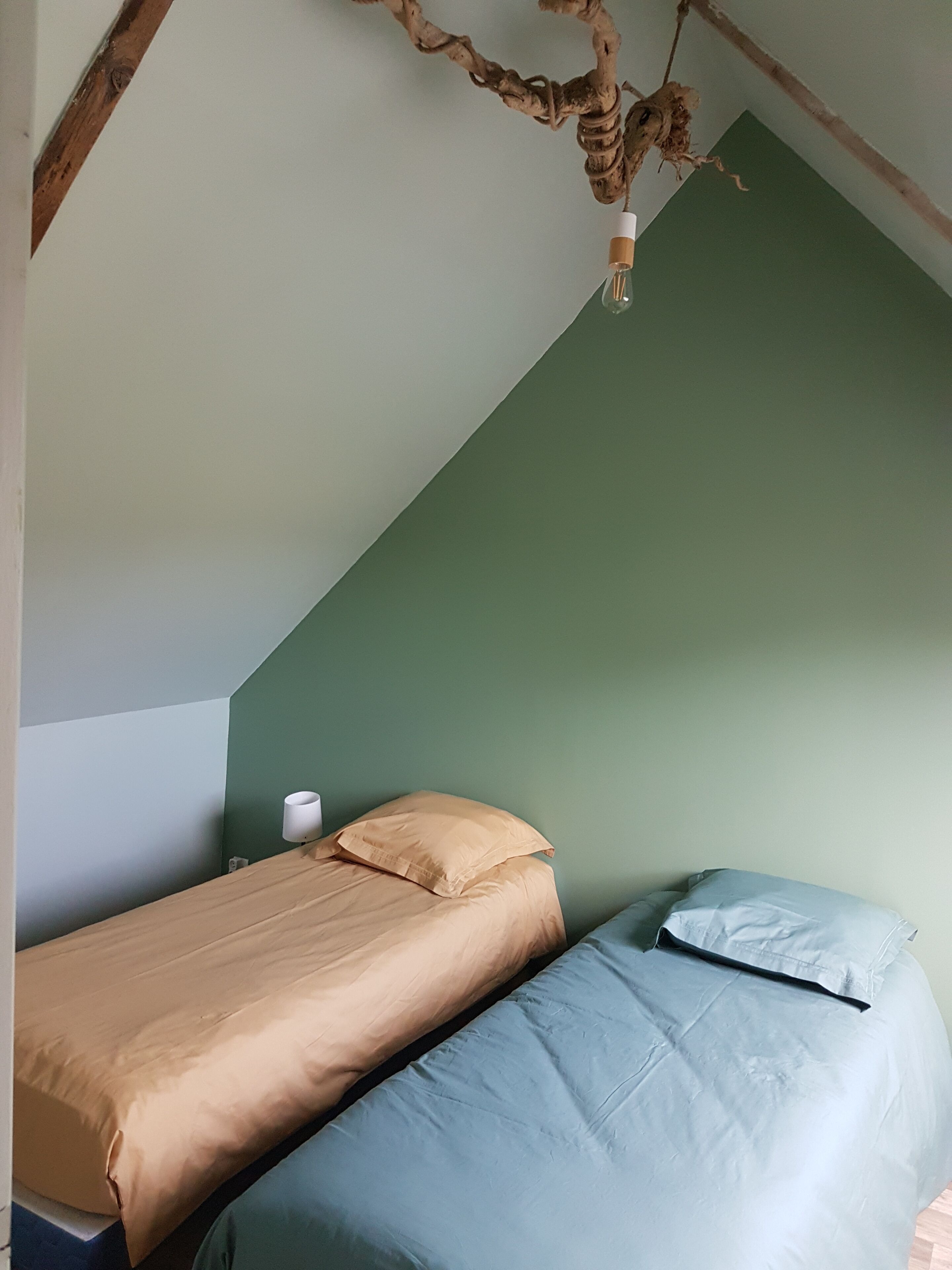 Ett litet, ombonat rum med ett sluttande tak, en säng med beige och blå sängkläder, en vit kopp på nattduksbordet och en rustik gren som hänger från taket.
