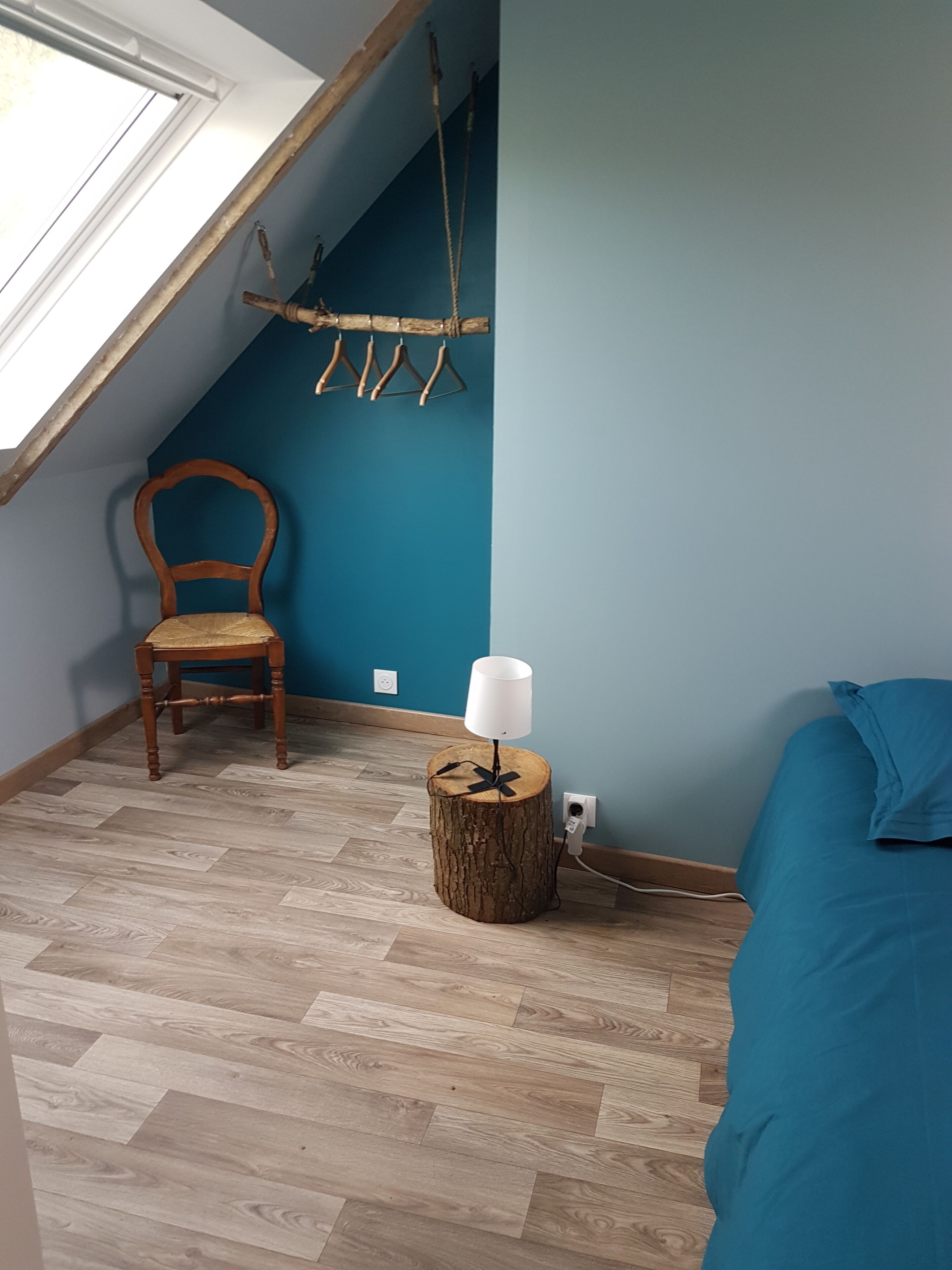 Une pièce avec un plafond en pente, une chaise en bois, une petite table en bois avec une lampe, un lit et un étendoir fixé au mur.