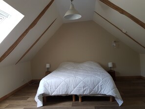 Ein Schlafzimmer mit geneigter Decke, einem Bett mit weißer Decke, zwei Nachttischlampen und einem Fenster, das natürliches Licht hereinlässt.