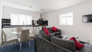 A modern living room with a grey sofa, red cushions, a glass coffee table, and a dining area with a white table and chairs. - Family Apartment 2nd Floor (Blackpool)