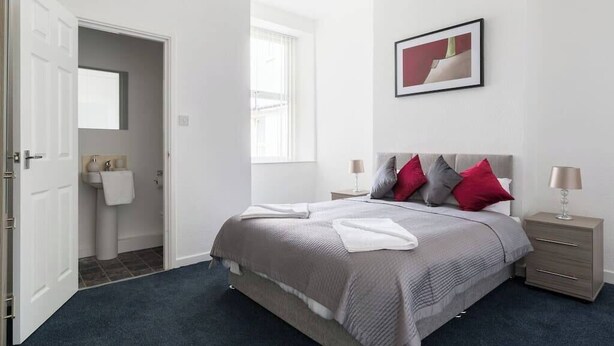 Une chambre à coucher comprenant un lit, des tables de chevet, une commode, une fenêtre, une porte et une salle de bain visible à travers une porte ouverte.