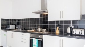 A modern kitchen with white cabinets, a black tiled backsplash, stainless steel appliances, and a built-in oven. - Two-Bedroom Apartment Ground Floor (Blackpool)