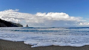A beach with waves, a rocky cliff, and a lighthouse in the distance.