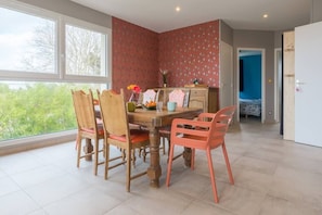Un comedor con mesa y sillas de madera, vistas a través de amplios ventanales y una pared con estampado rojo.