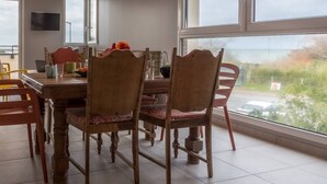 A dining area with a wooden table and chairs, a TV mounted on the wall, and a view of the outdoors through large windows. - Tribord, apartment with terrace and sea view, parking (Saint-Côme-de-Fresné)