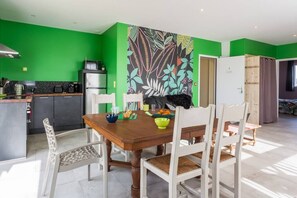 A dining area with a wooden table and white chairs, a green wall with a floral mural, and a kitchen in the background.