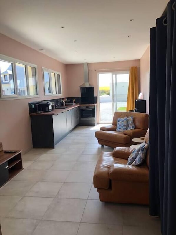 A modern living room with a brown leather sofa, a kitchen area with a stove and microwave, and a large window with curtains.