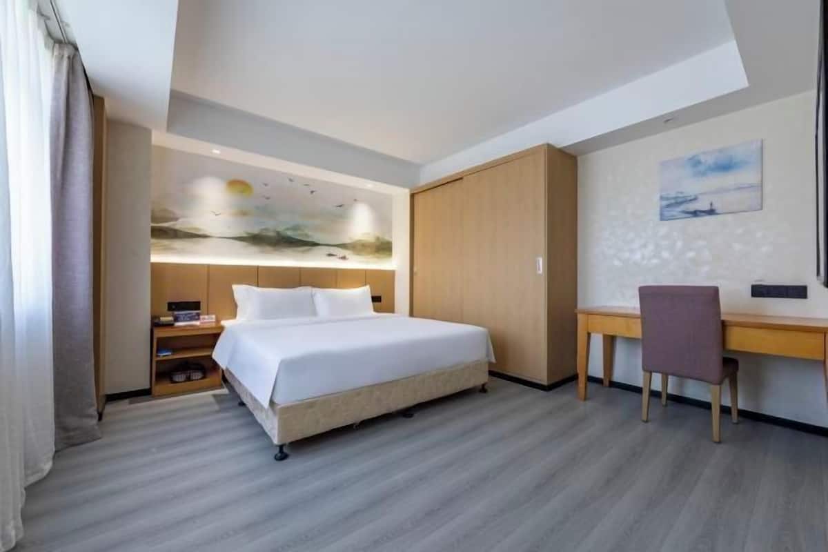 A hotel room with a bed, a desk, and a chair. There is a painting on the wall and a window with curtains.