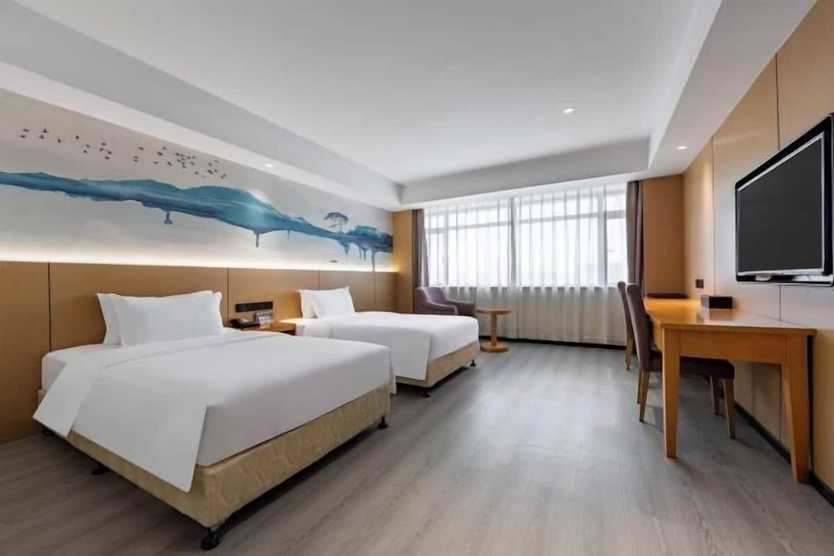 A hotel room with two beds, a desk, a chair, a TV, and a large window with curtains.