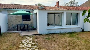 Una casa pequeña con techo de tejas, pared de piedra y una ventana. Hay una mesa y sillas afuera.
