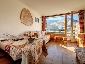 A cozy living room with a sofa, dining table, and chairs, a large window with a mountain view, and a woven basket on the wall. - Bright studio in Menuires, ski-in/ski-out, balcony (Les Menuires)