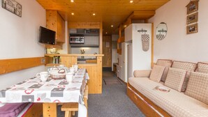 A cozy room with a wooden ceiling, a dining table with a patterned tablecloth, a sofa with patterned cushions, a television on the wall, and a kitchen area in the background. - Renovated duplex in quiet area with balcony - 6/7 pers. (Les Arcs 1800)
