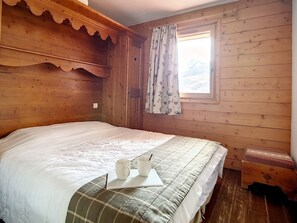 A wooden cabin bedroom with a bed, a window with curtains, and a small table with two mugs. - Spacious apartment near slopes with balcony and parking (Les Menuires)