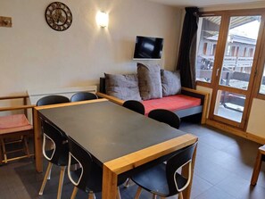 Quarto com mesa de madeira, cadeiras pretas, sofá com almofadas, televisão na parede e janela com cortinas.