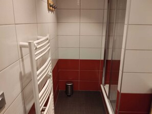 Un baño con baldosas blancas y rojas, un perchero blanco, un cesto de basura y un box de ducha de vidrio.